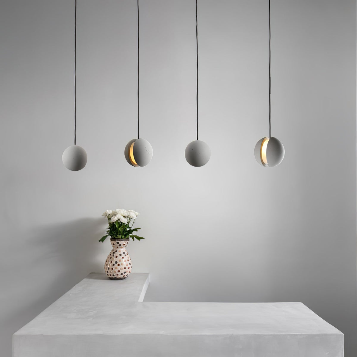 Four Clérachic spheres hang in a row over a pale concrete counter, with a spotted vase of white blooms beneath them.