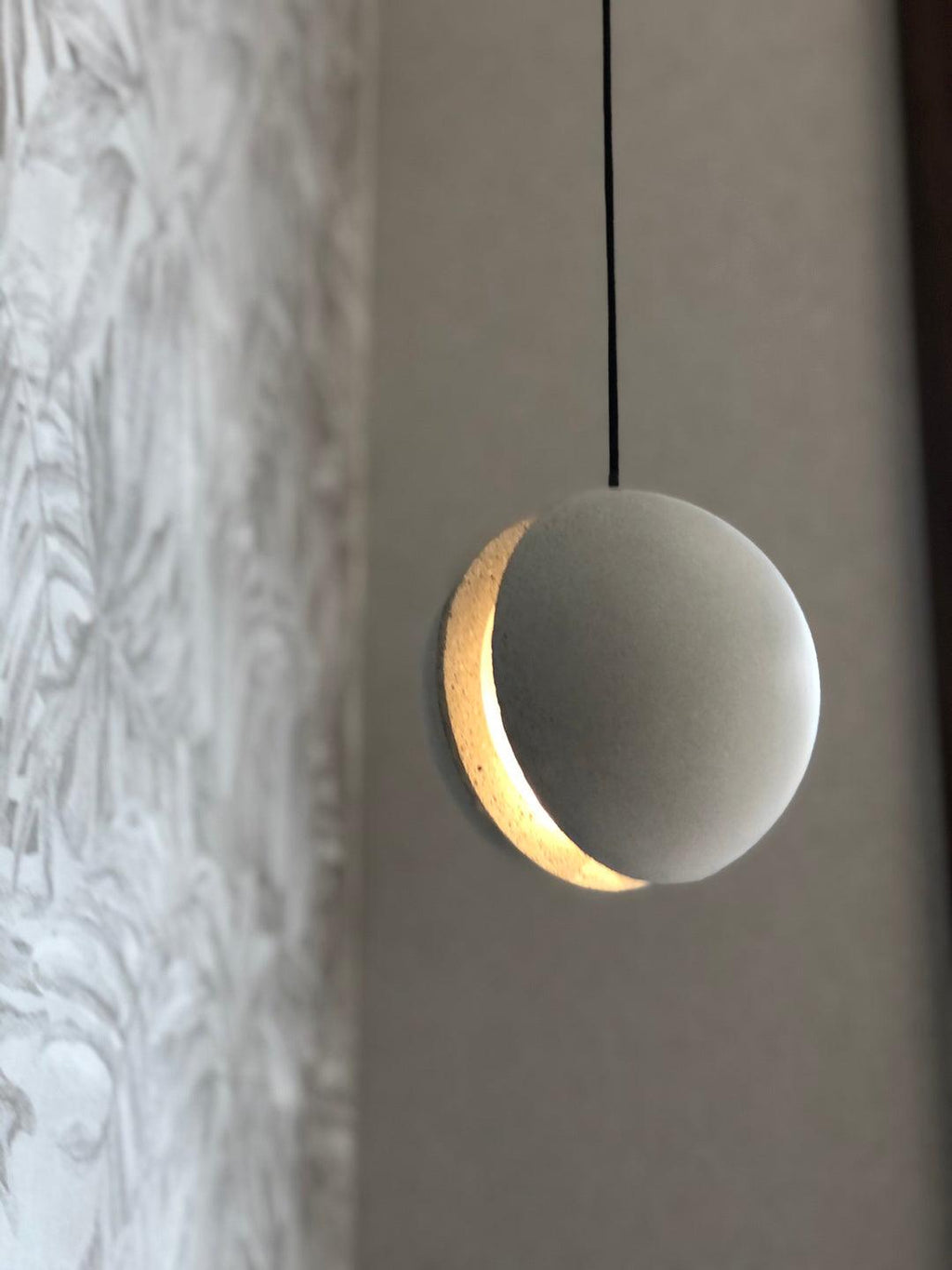 Close view of a Clérachic concrete pendant, its crescent slot glowing warm against a pale mottled wall.
