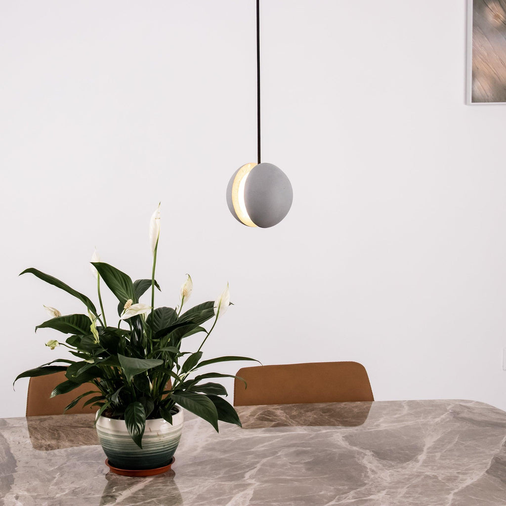 A single Clérachic concrete pendant with a lit slot hangs above a marble dining table and a leafy white-flowered plant.