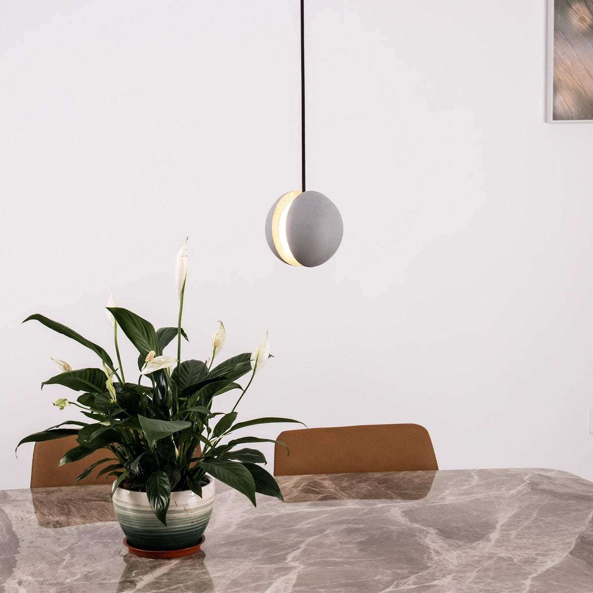 A single Clérachic concrete pendant with a lit slot hangs above a marble dining table and a leafy white-flowered plant.