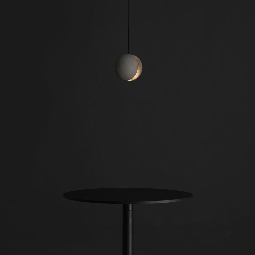 A small Clérachic concrete pendant glows through its slot in a dark room, above a slim black round table.