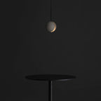 A small Clérachic concrete pendant glows through its slot in a dark room, above a slim black round table.