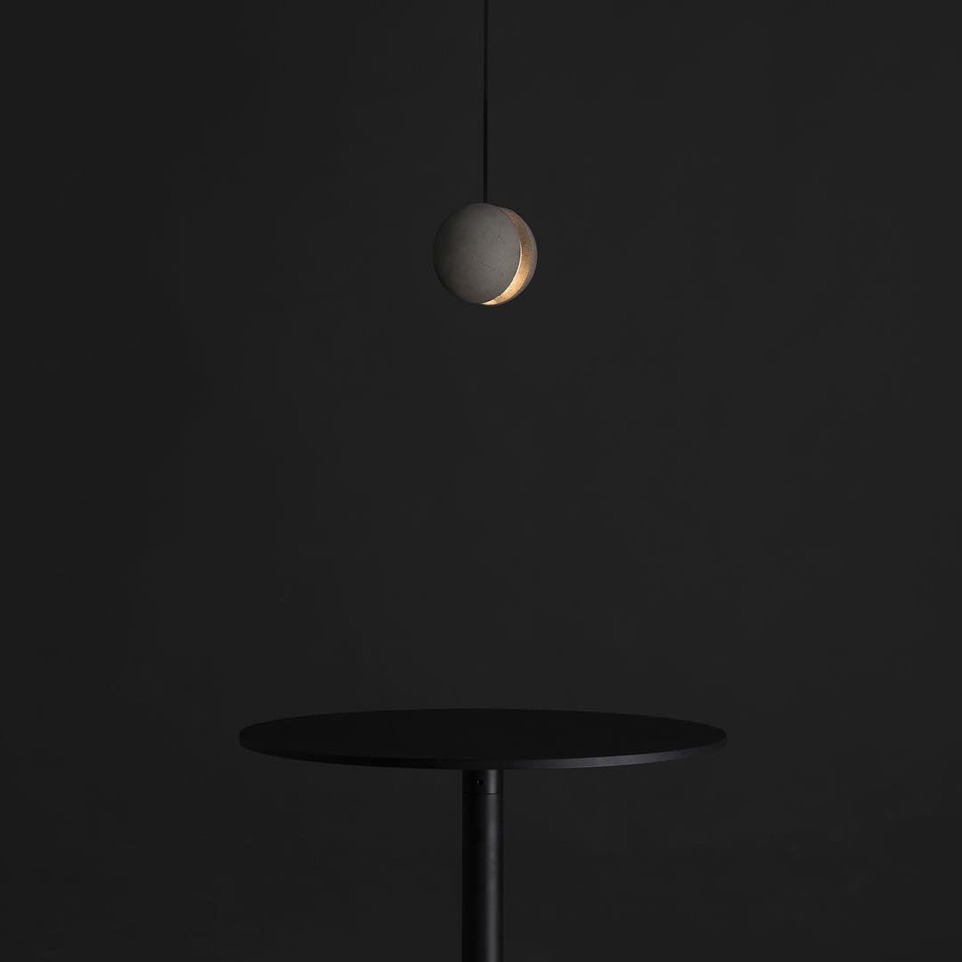 A small Clérachic concrete pendant glows through its slot in a dark room, above a slim black round table.