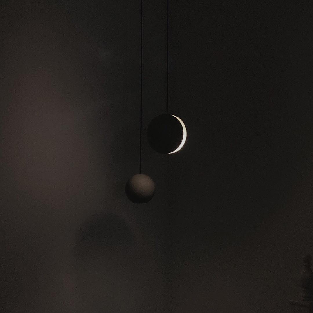Two Clérachic concrete pendants hang in near darkness, one showing a thin bright crescent of light.