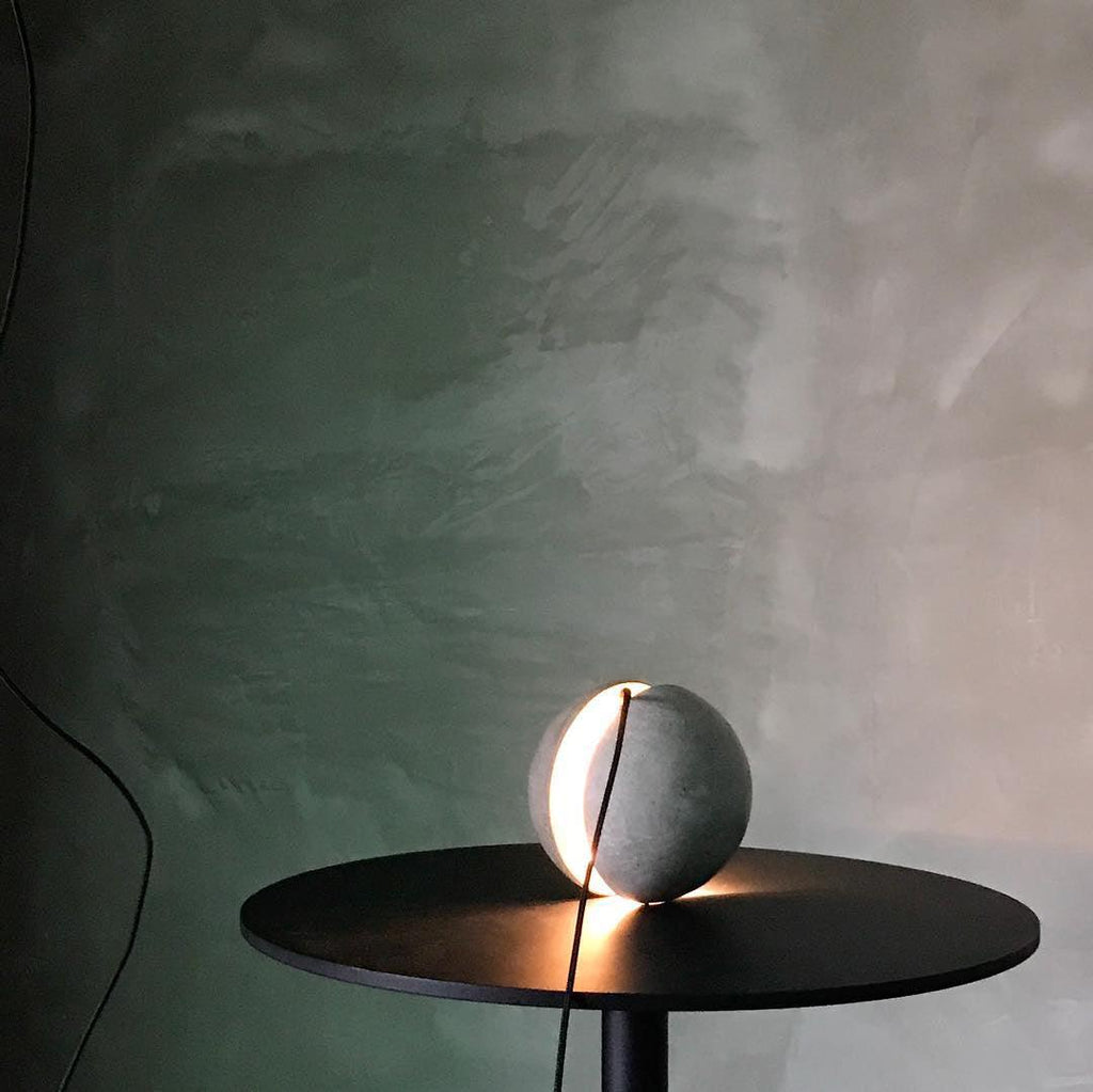 A Clérachic concrete sphere rests on a black round table with its slot glowing, against a green-tinted plaster wall.
