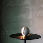 A Clérachic concrete sphere rests on a black round table with its slot glowing, against a green-tinted plaster wall.
