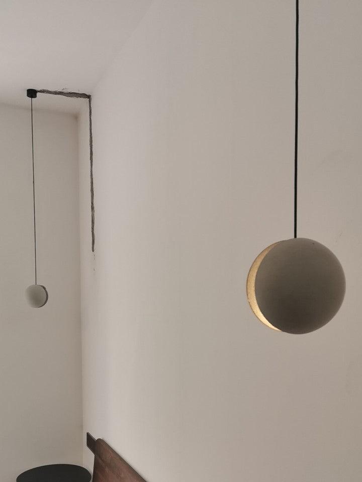 Two Clérachic pendants hang from a white wall, the nearer one showing a warm crescent slot, with a dark stool below.