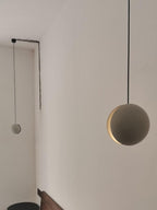 Two Clérachic pendants hang from a white wall, the nearer one showing a warm crescent slot, with a dark stool below.
