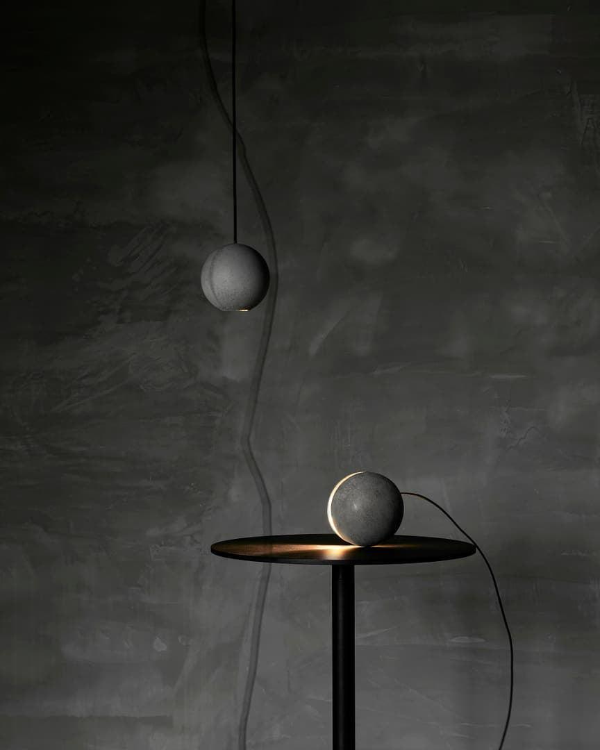 One Clérachic concrete sphere hangs above a dark table where a second sits lit, against a dark plaster wall.