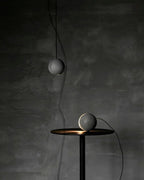 One Clérachic concrete sphere hangs above a dark table where a second sits lit, against a dark plaster wall.