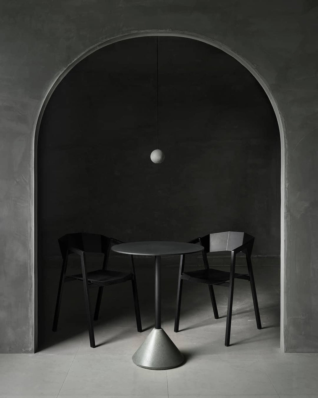 A tiny Clérachic sphere hangs inside a stone archway over a small round table and two black chairs on a grey floor.