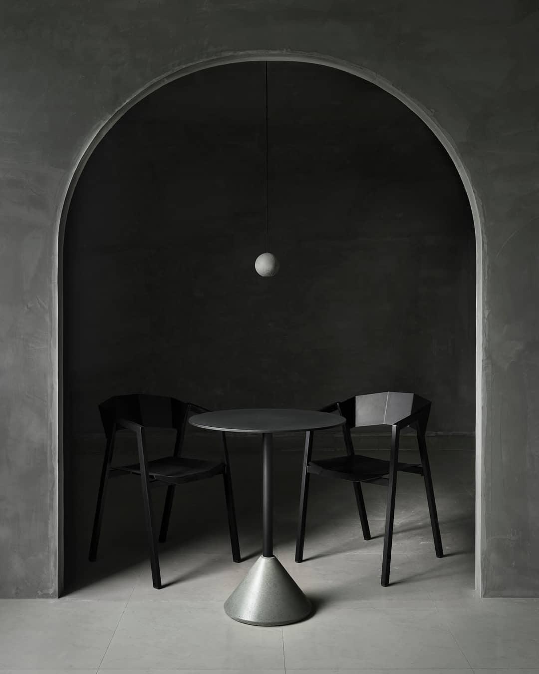 A tiny Clérachic sphere hangs inside a stone archway over a small round table and two black chairs on a grey floor.