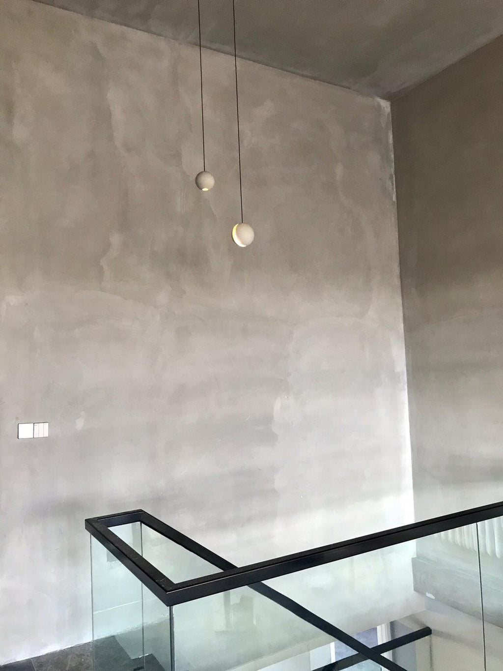 Two small Clérachic pendants hang against a tall plaster wall above a stairwell with a glass and black-metal rail.