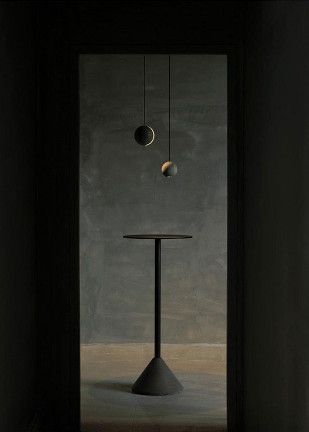 Two Clérachic concrete pendants float in a dark doorway above a tall black pedestal table on a cone base.