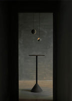 Two Clérachic concrete pendants float in a dark doorway above a tall black pedestal table on a cone base.