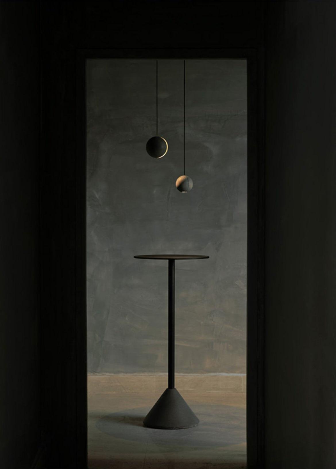 Two Clérachic concrete pendants float in a dark doorway above a tall black pedestal table on a cone base.