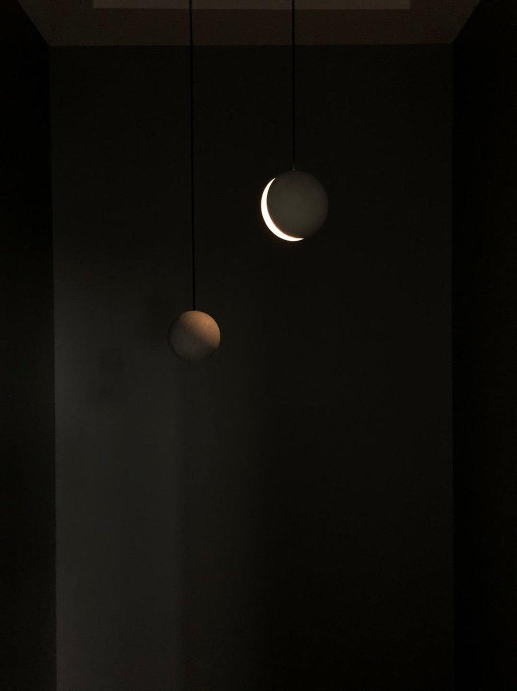 Two Clérachic pendants glow in a dark room, one showing a bright crescent of light and the other a dim warm edge.