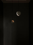 Two Clérachic pendants glow in a dark room, one showing a bright crescent of light and the other a dim warm edge.