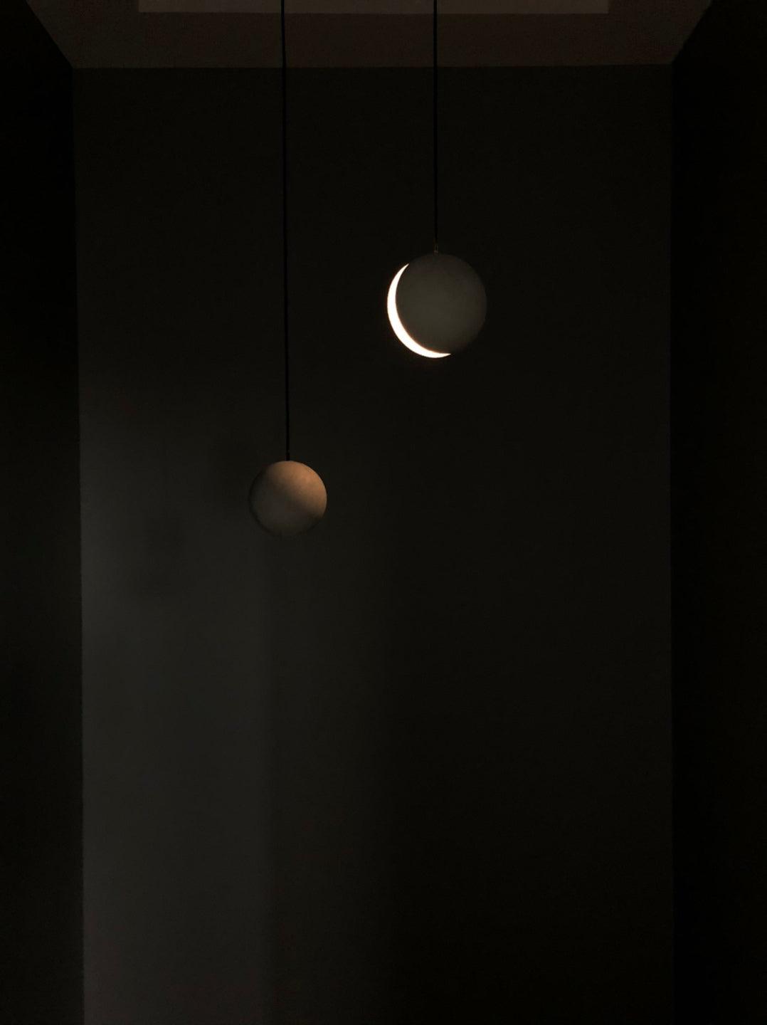 Two Clérachic pendants glow in a dark room, one showing a bright crescent of light and the other a dim warm edge.