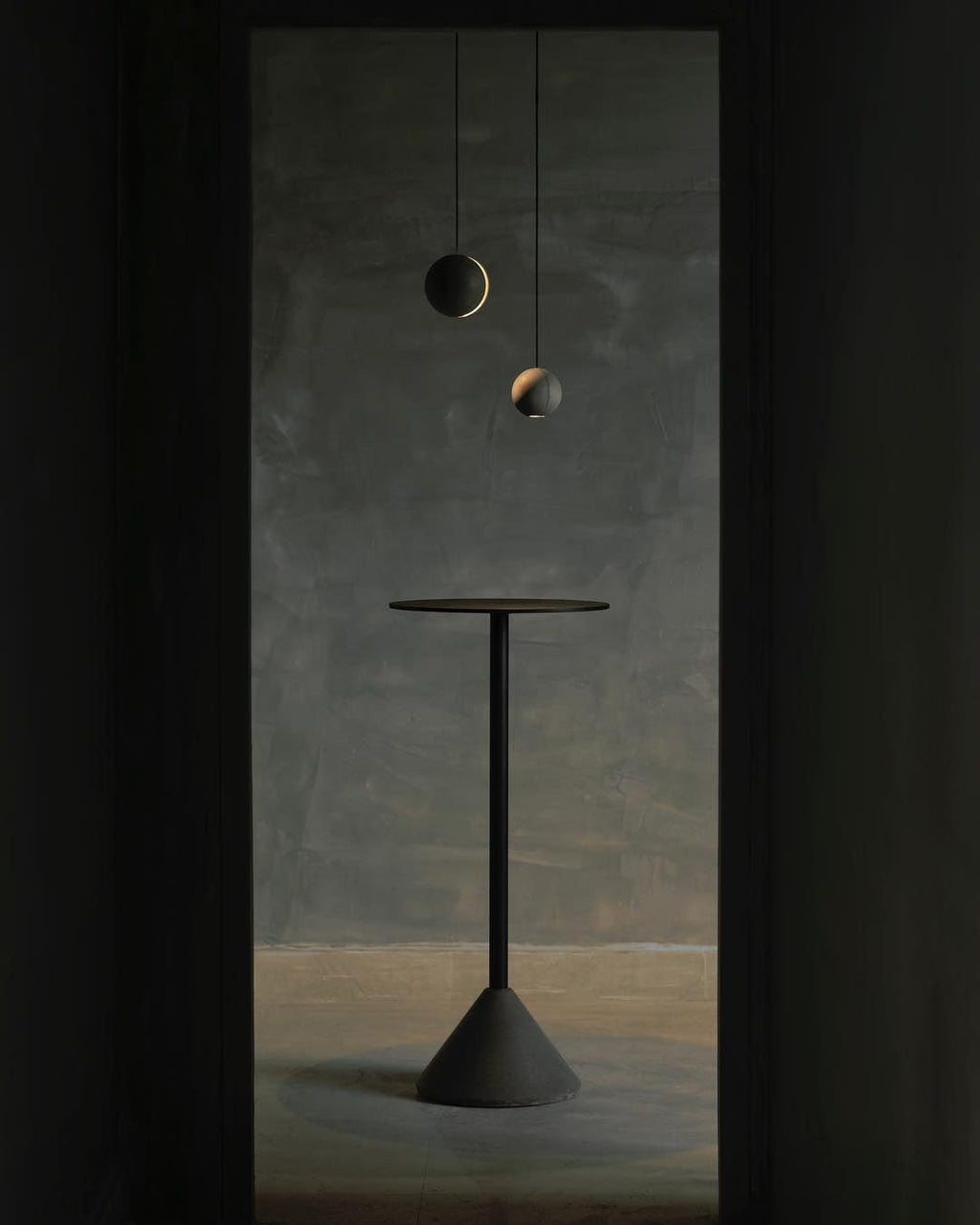 Two Clérachic concrete pendants hang in a dark doorway, one with a visible crescent glow, over a tall dark table.
