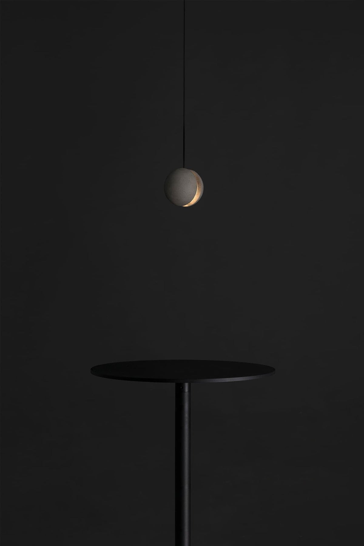 A single Clérachic concrete pendant with a soft crescent glow hangs over a slim black table against a dark wall.