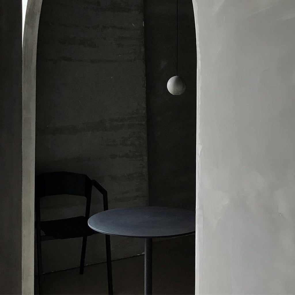 A cement-grey Clérachic sphere hangs in a dim corner beside a curved plaster wall, over a round table and a black chair.