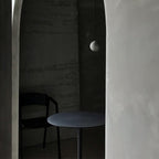 A cement-grey Clérachic sphere hangs in a dim corner beside a curved plaster wall, over a round table and a black chair.