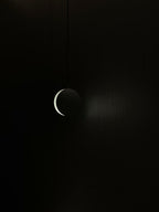 A Clérachic concrete pendant seen in near darkness, with only a thin curve of light along its slot.