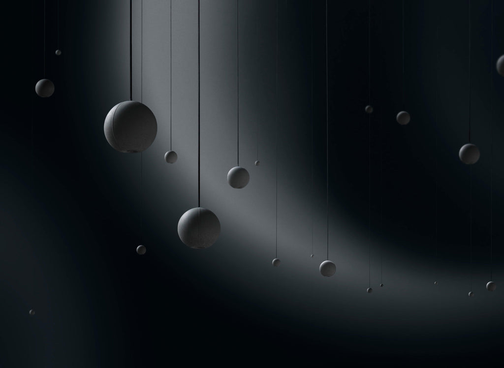 A scatter of Clérachic concrete spheres of many sizes hangs on fine cords in a dark space, lit by a sweeping beam.