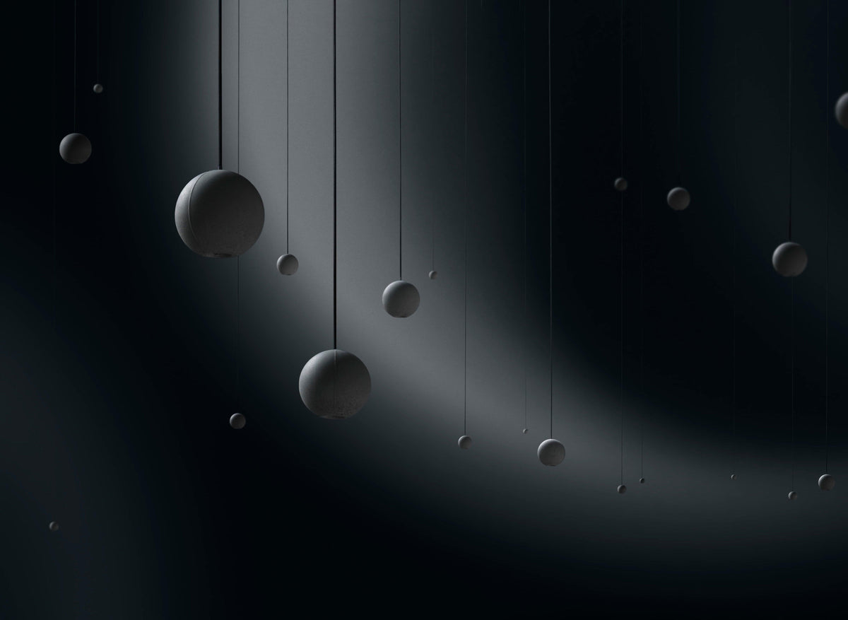 A scatter of Clérachic concrete spheres of many sizes hangs on fine cords in a dark space, lit by a sweeping beam.
