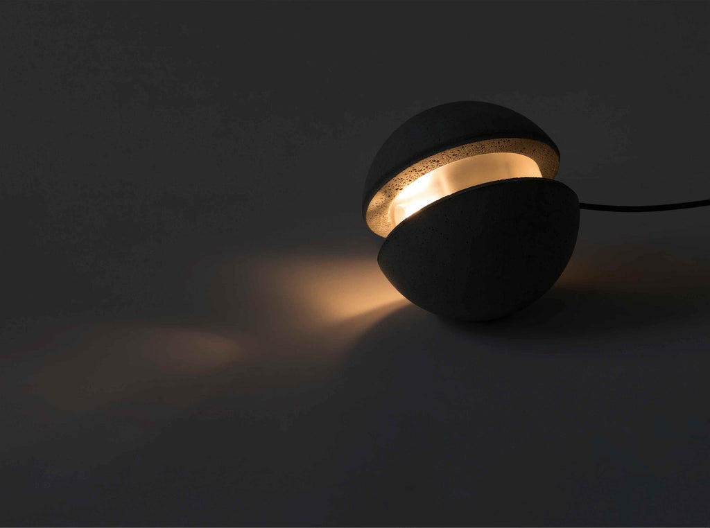 A Clérachic concrete sphere with a glowing slot sits on a dark floor, its warm light spilling across the surface.