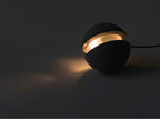 A Clérachic concrete sphere with a glowing slot sits on a dark floor, its warm light spilling across the surface.