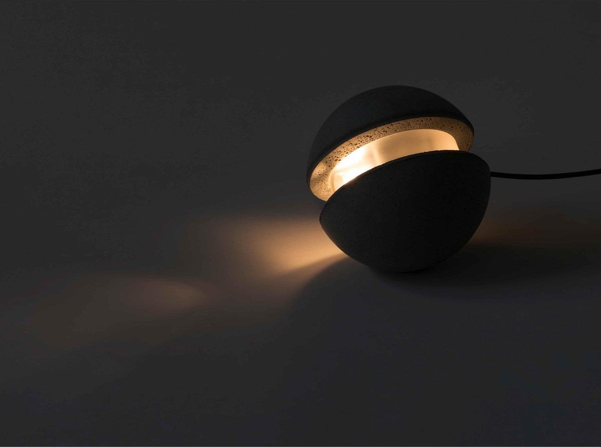 A Clérachic concrete sphere with a glowing slot sits on a dark floor, its warm light spilling across the surface.