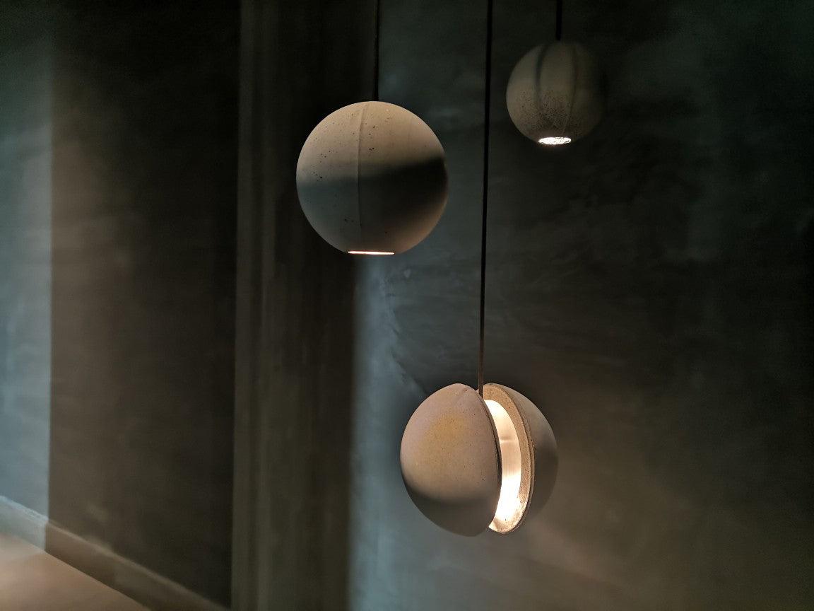 Three Clérachic concrete pendants hang against a dark wall, the lowest glowing warmly through its crescent slot.
