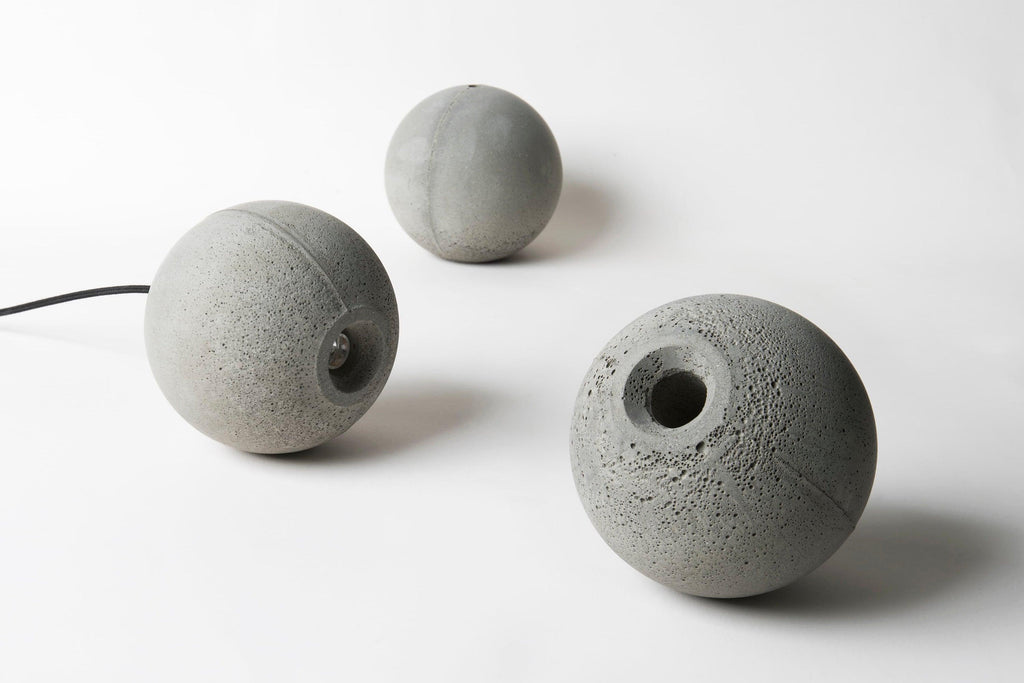 Three unlit Clérachic concrete spheres lie on a white surface, one with a round hole and a black cord trailing out.