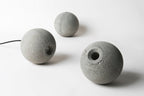 Three unlit Clérachic concrete spheres lie on a white surface, one with a round hole and a black cord trailing out.