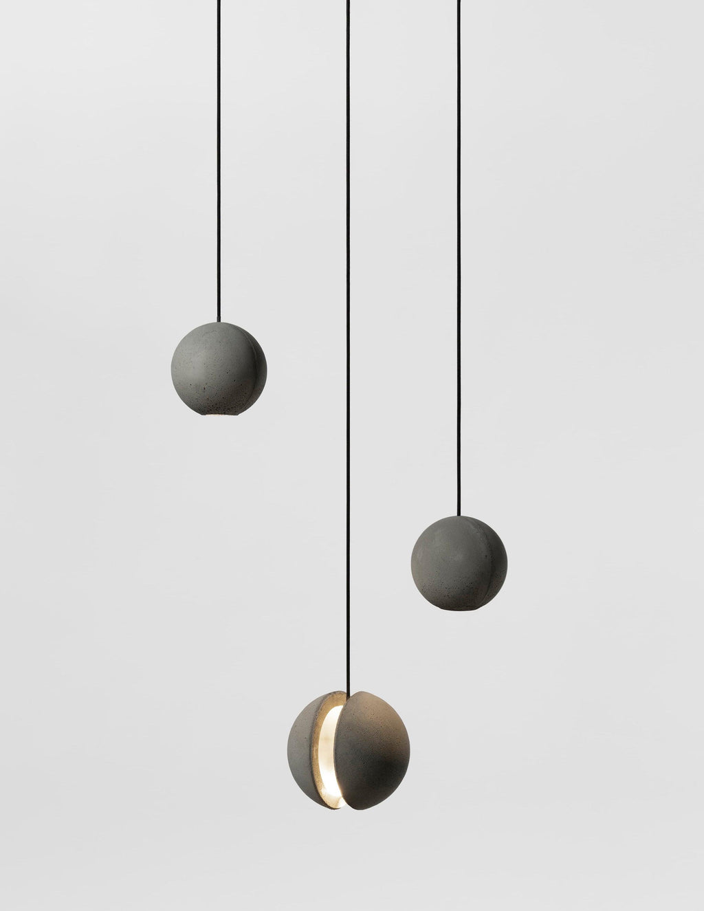Three smaller Clérachic concrete pendants hang on black cords in a grey studio, the lowest lit through its slot.
