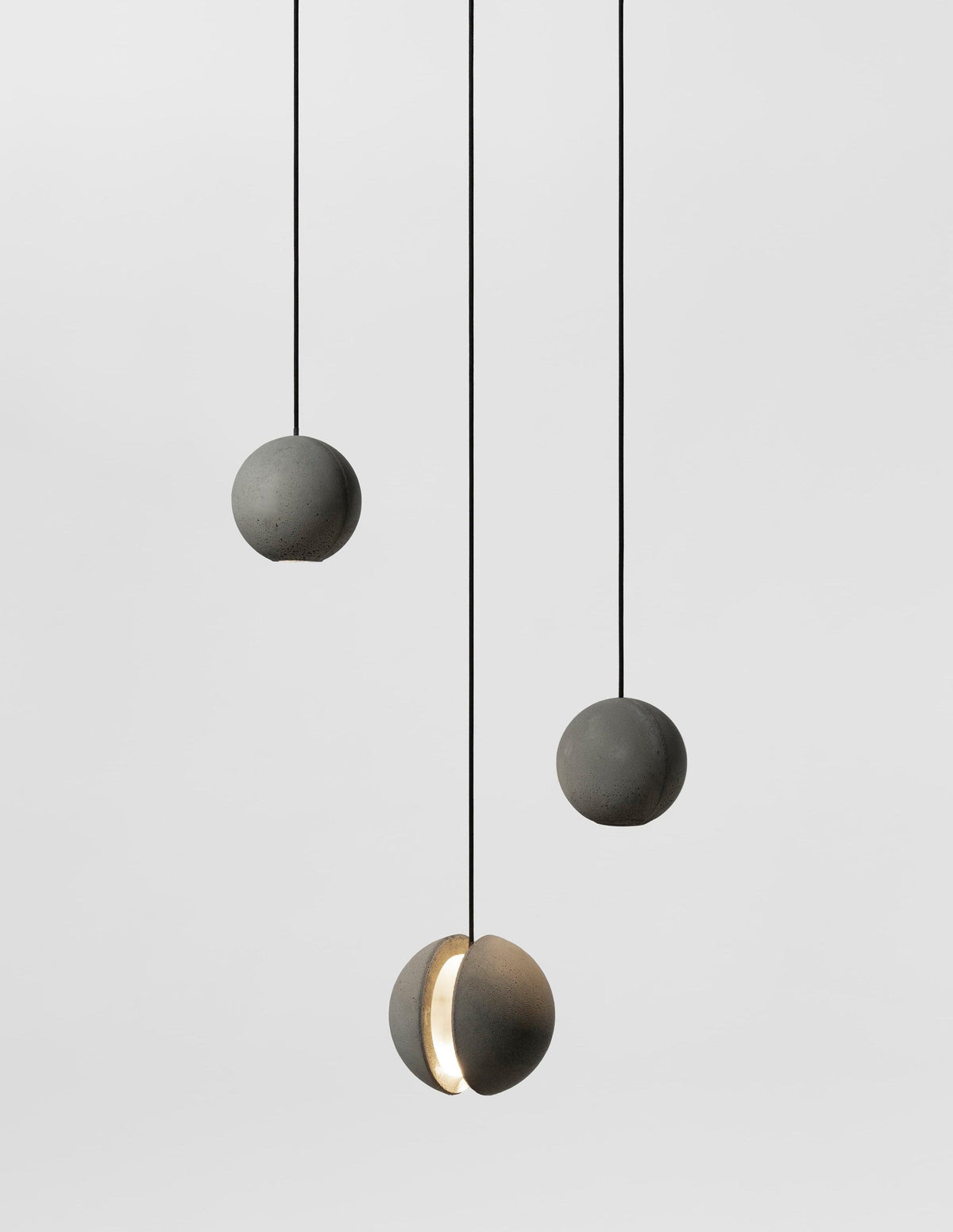 Three smaller Clérachic concrete pendants hang on black cords in a grey studio, the lowest lit through its slot.