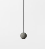 A single plain cement-grey Clérachic sphere hangs on a thin black cord against a pale backdrop.