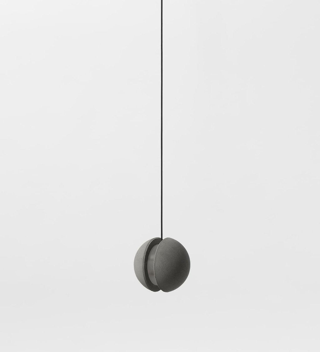 A Clérachic concrete pendant with a closed crescent slot hangs on a black cord against a pale backdrop, unlit.