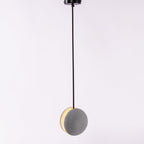 A Clérachic concrete pendant hangs from a black canopy on a lilac-grey backdrop, its crescent slot glowing warm.