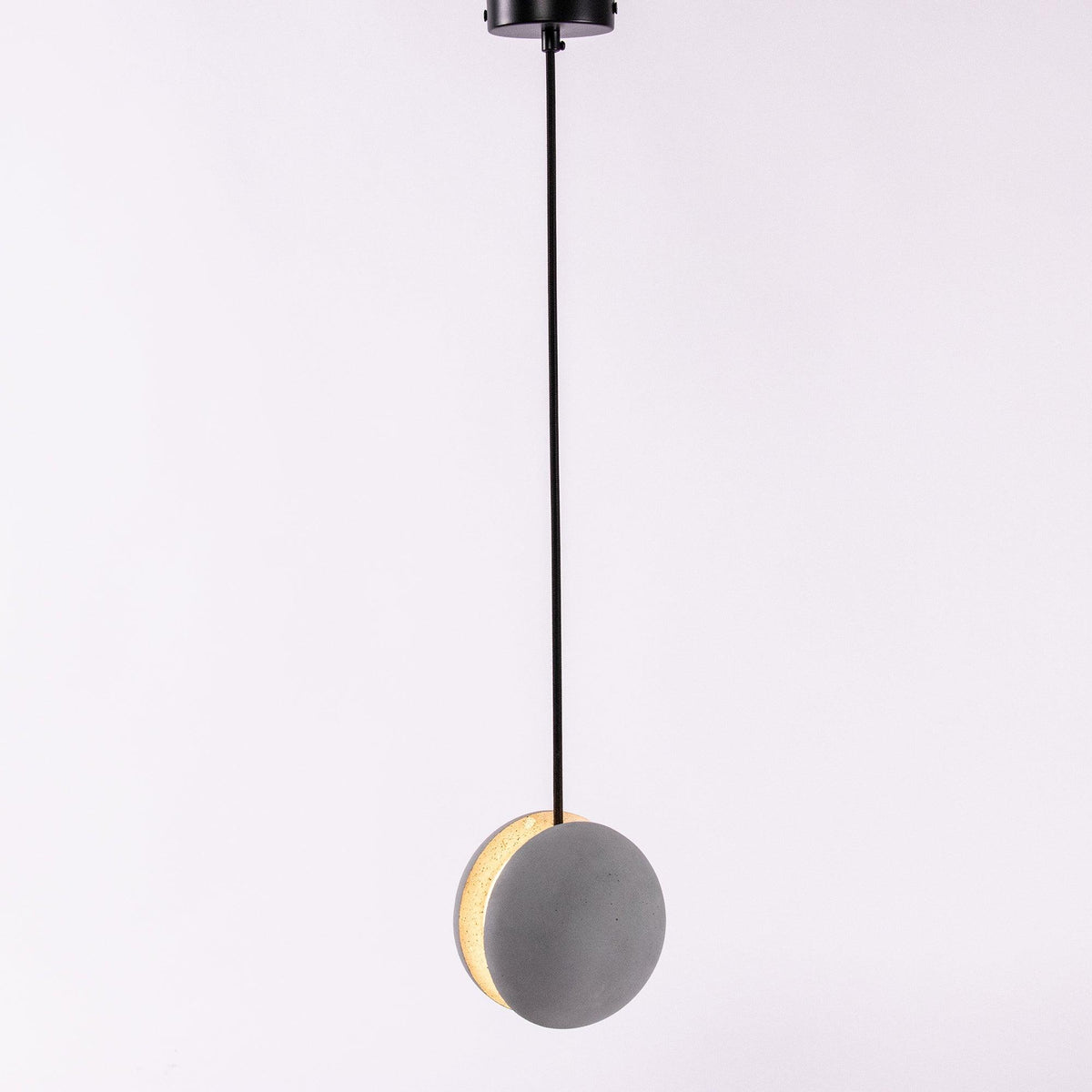 A Clérachic concrete pendant hangs from a black canopy on a lilac-grey backdrop, its crescent slot glowing warm.