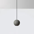 A cement-grey Clérachic sphere pendant hangs on a black cord against a pale grey wall and white floor.