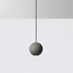 A cement-grey Clérachic sphere pendant hangs on a black cord against a pale grey wall and white floor.