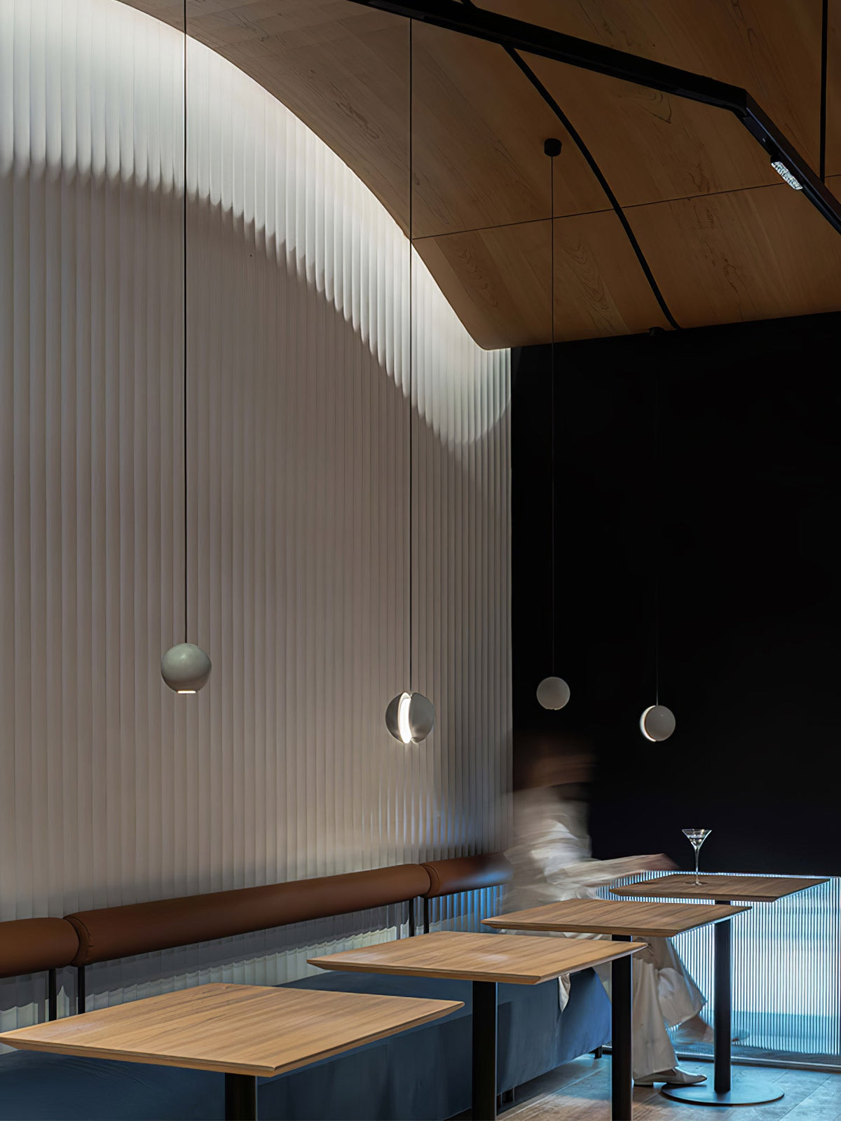 Small Clérachic pendants hang in a bar with a ribbed curved wall, a timber ceiling and wooden tables below.