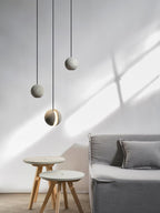 Three Clérachic concrete pendants hang at staggered heights in sunlight, above two wooden-legged stools and a grey sofa.