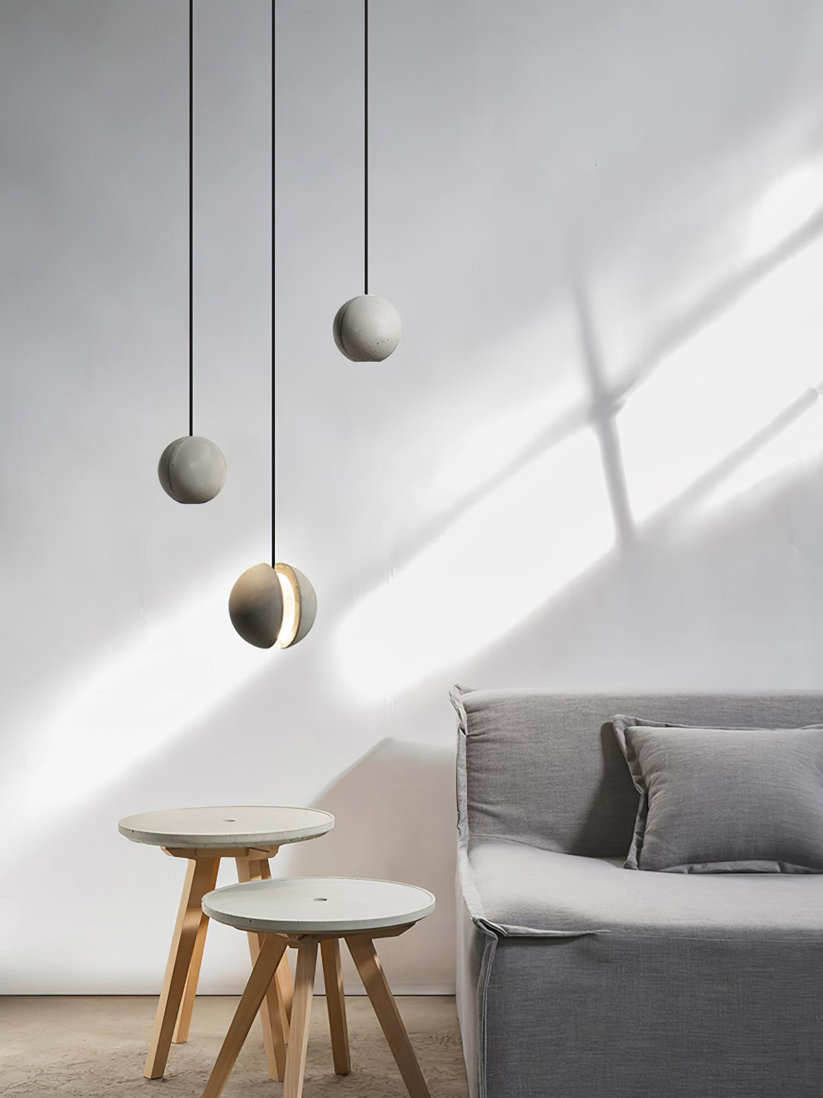 Three Clérachic concrete pendants hang at staggered heights in sunlight, above two wooden-legged stools and a grey sofa.