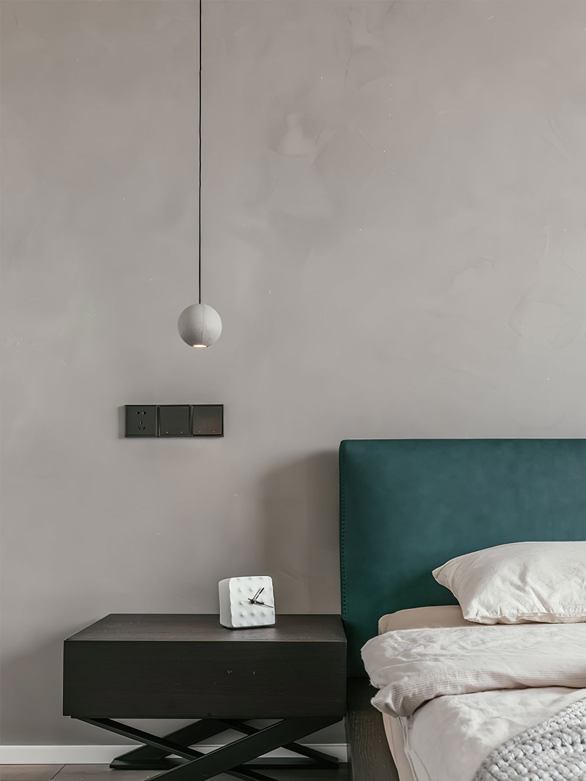 A small Clérachic concrete pendant hangs by a bedside, beside a dark nightstand and a green headboard.