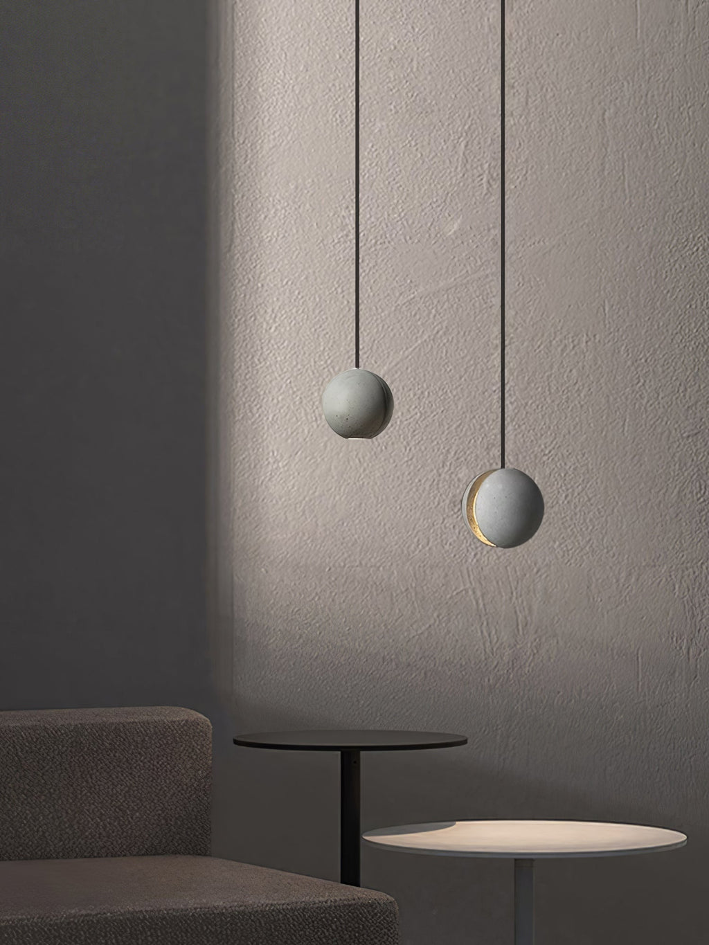 Two Clérachic concrete pendants hang against a softly lit plaster wall, the lower one lit, above a dark side table.