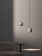 Two Clérachic concrete pendants hang against a softly lit plaster wall, the lower one lit, above a dark side table.